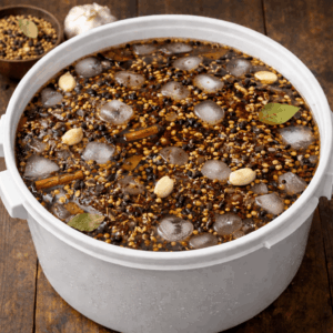 Large white pot filled with a dark, spiced stew or broth dotted with ice cubes, seeds, and nuts, resting on a wooden surface.