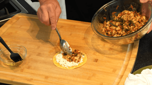 Person assembles a taco by spooning seasoned filling onto a small tortilla on a wooden board, with a bowl of filling nearby.
