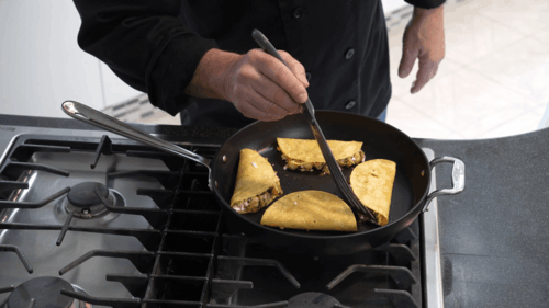 Person cooking folded tortillas in a black skillet on a gas stove, using tongs to flip them on a countertop.