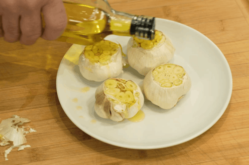 Person pours olive oil over roasted garlic bulbs arranged on a white plate in a kitchen setting.