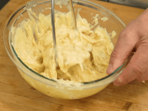 Hands beating creamy yellow batter in a glass bowl on a wooden counter.